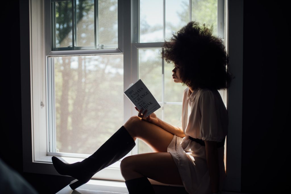 A silhouetted woman with afro hair reading a book by a window. Photo by Thought Catalog from Pexels